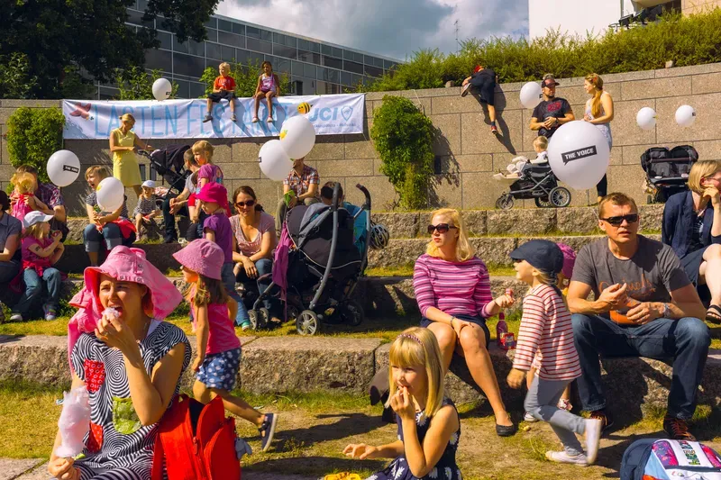 A crowd of families sits on stone steps at an outdoor children's festival, a woman in a pink sunhat eating cotton candy in the foreground and white balloons and strollers behind them.
