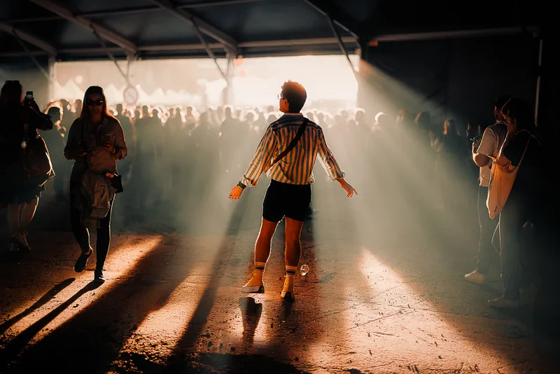 A man in a striped shirt dances alone in a shaft of golden light inside a festival tent, a crowd silhouetted in the haze behind him.