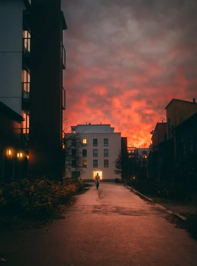 A figure walks toward a glowing lit doorway between apartment buildings beneath a fiery orange and pink sunset sky.