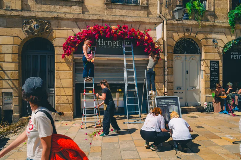 Florists on ladders build a cascading arch of pink and red blooms over a shopfront while passersby move through the sunny square.