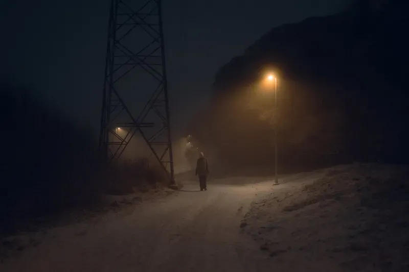 A person in a white beanie walks a snowy path past an electricity pylon, a single streetlamp glowing in the fog.