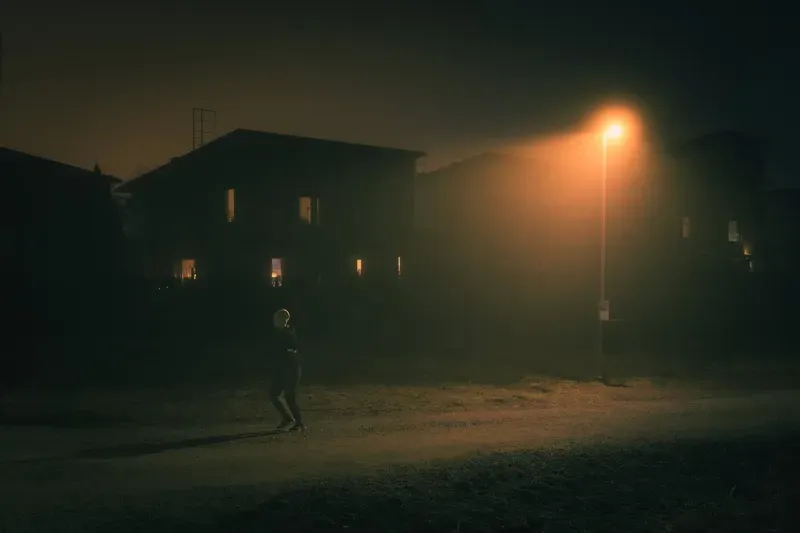 A person walks in silhouette past an apartment building whose lit windows glow through the fog.