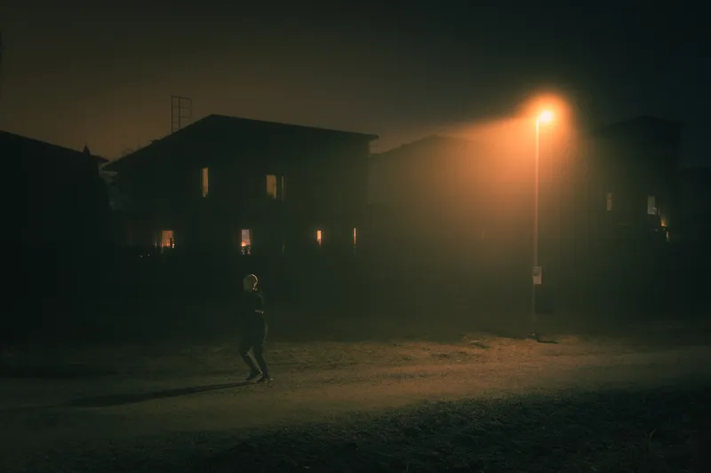 A person walks in silhouette past an apartment building whose lit windows glow through the fog.
