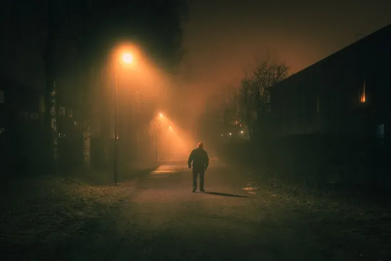 A man in a red beanie walks away down a misty street lit only by rows of orange streetlights.