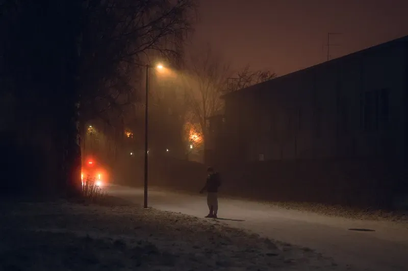 A person with a backpack walks along a foggy street past red car taillights and a dark apartment building.