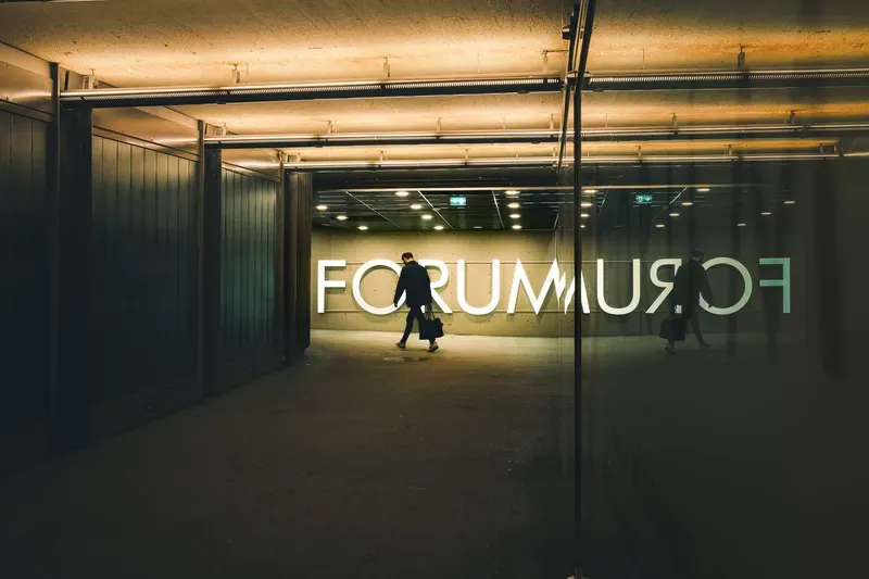 A man carrying a bag walks through an illuminated underpass beneath a giant FORUM sign, his reflection mirrored in the glass beside him.