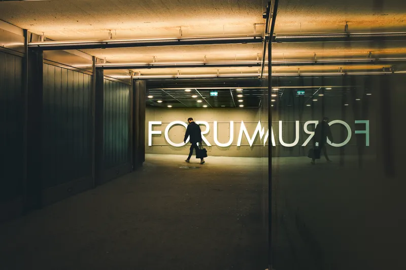 A man carrying a bag walks through an illuminated underpass beneath a giant FORUM sign, his reflection mirrored in the glass beside him.