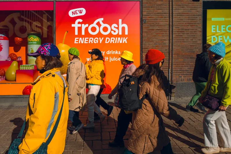 A cluster of colorfully dressed pedestrians in caps and beanies walks past a bright orange Froosh energy drink billboard.