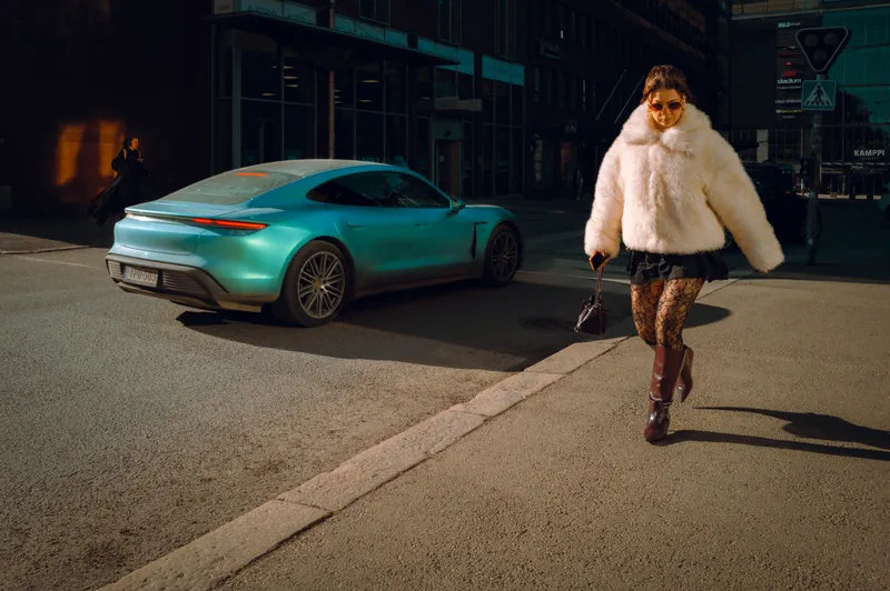 A woman in a white fur coat and sunglasses crosses a street at night past a parked teal sports car and glowing shopfronts.