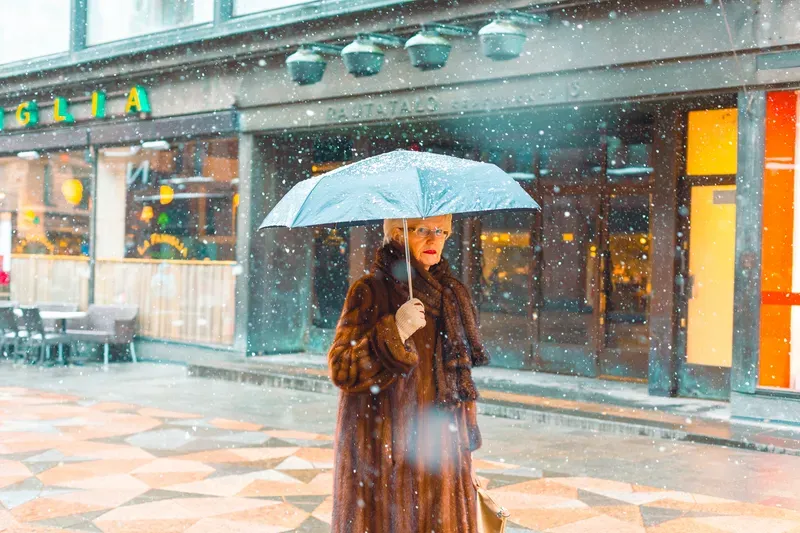 An elderly woman in a fur coat holds a pale blue umbrella against falling snow outside a row of lit shopfronts.