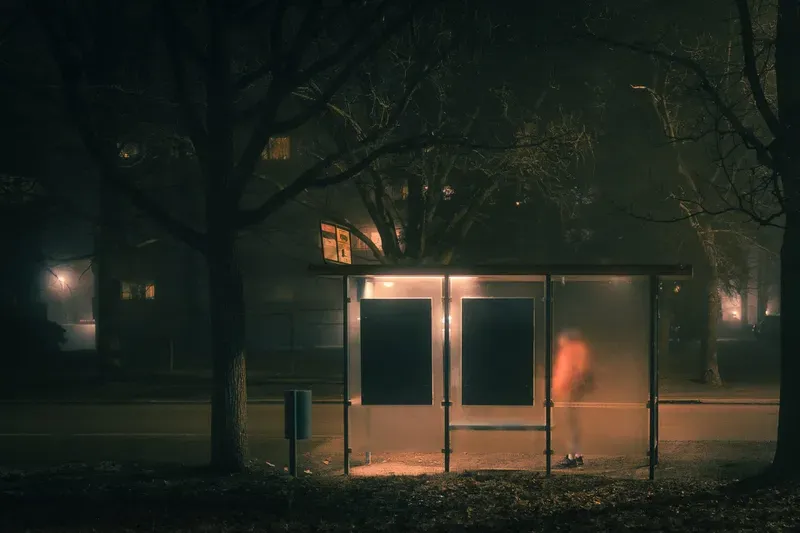 A bus shelter glows amber in the fog, a blurred figure in orange visible through its frosted glass panel.