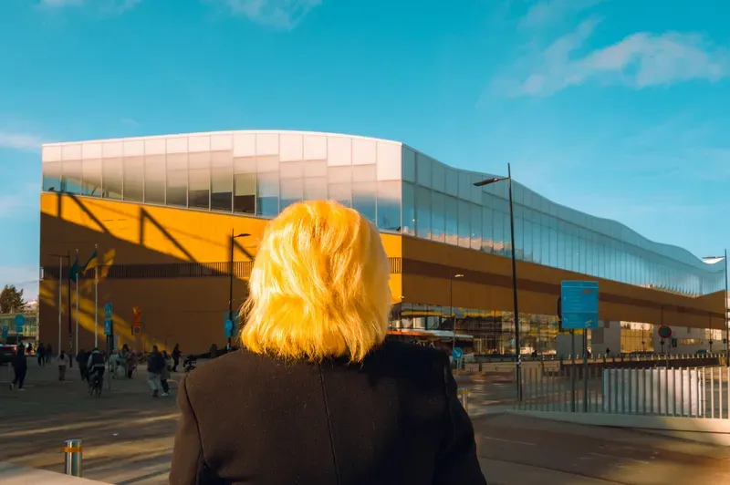 Seen from behind, a woman with sunlit blonde hair looks toward the curved wooden and glass facade of a modern library building.