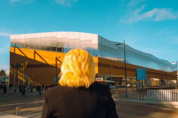 Seen from behind, a woman with sunlit blonde hair looks toward the curved wooden and glass facade of a modern library building.
