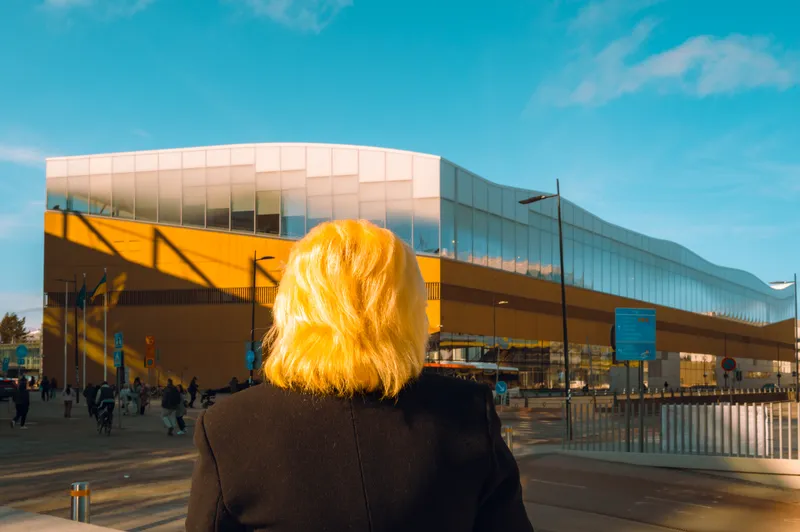 Seen from behind, a woman with sunlit blonde hair looks toward the curved wooden and glass facade of a modern library building.