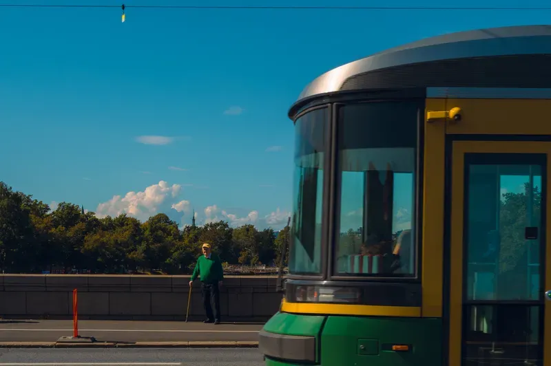 A green and yellow tram stands in bright sunlight under a deep blue sky, a man in green leaning on a walking stick on the pavement beyond.