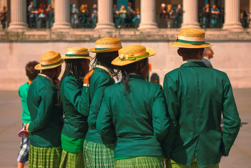 A line of schoolchildren in green blazers and straw boater hats stand before a colonnaded building.