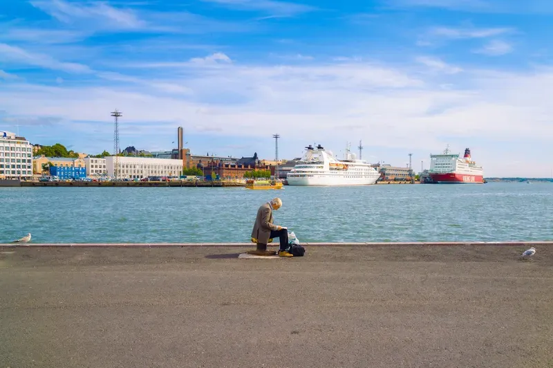 An elderly man sits reading on a harbor bollard beside gulls, with cruise ships and a Viking Line ferry docked across the water.