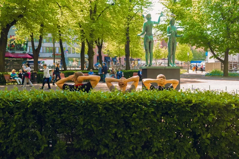 Three men sit in a row behind a hedge, hands clasped behind their heads, facing a bronze statue of two figures in a sunlit park.