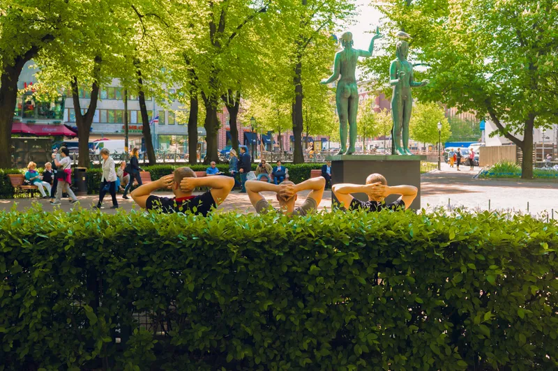 Three men sit in a row behind a hedge, hands clasped behind their heads, facing a bronze statue of two figures in a sunlit park.
