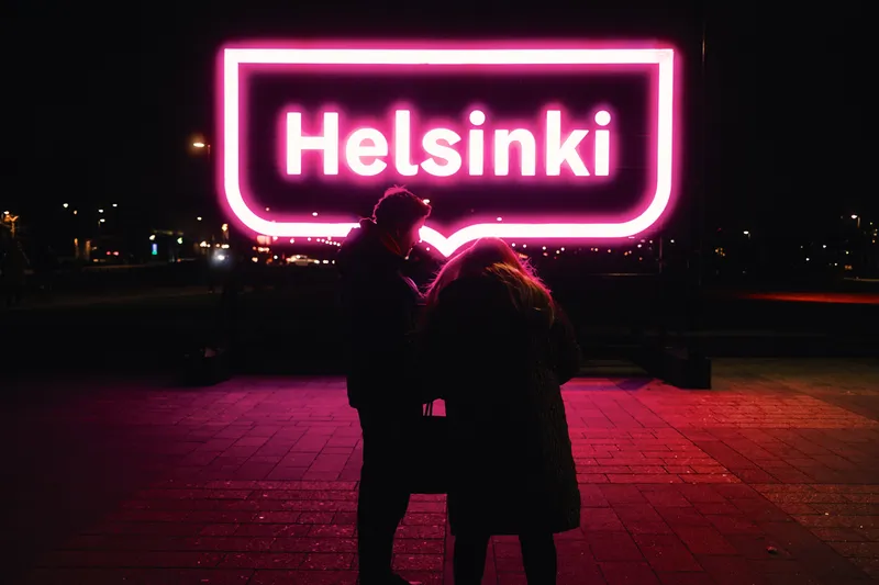 A couple embraces at night beneath a glowing pink neon sign reading Helsinki.