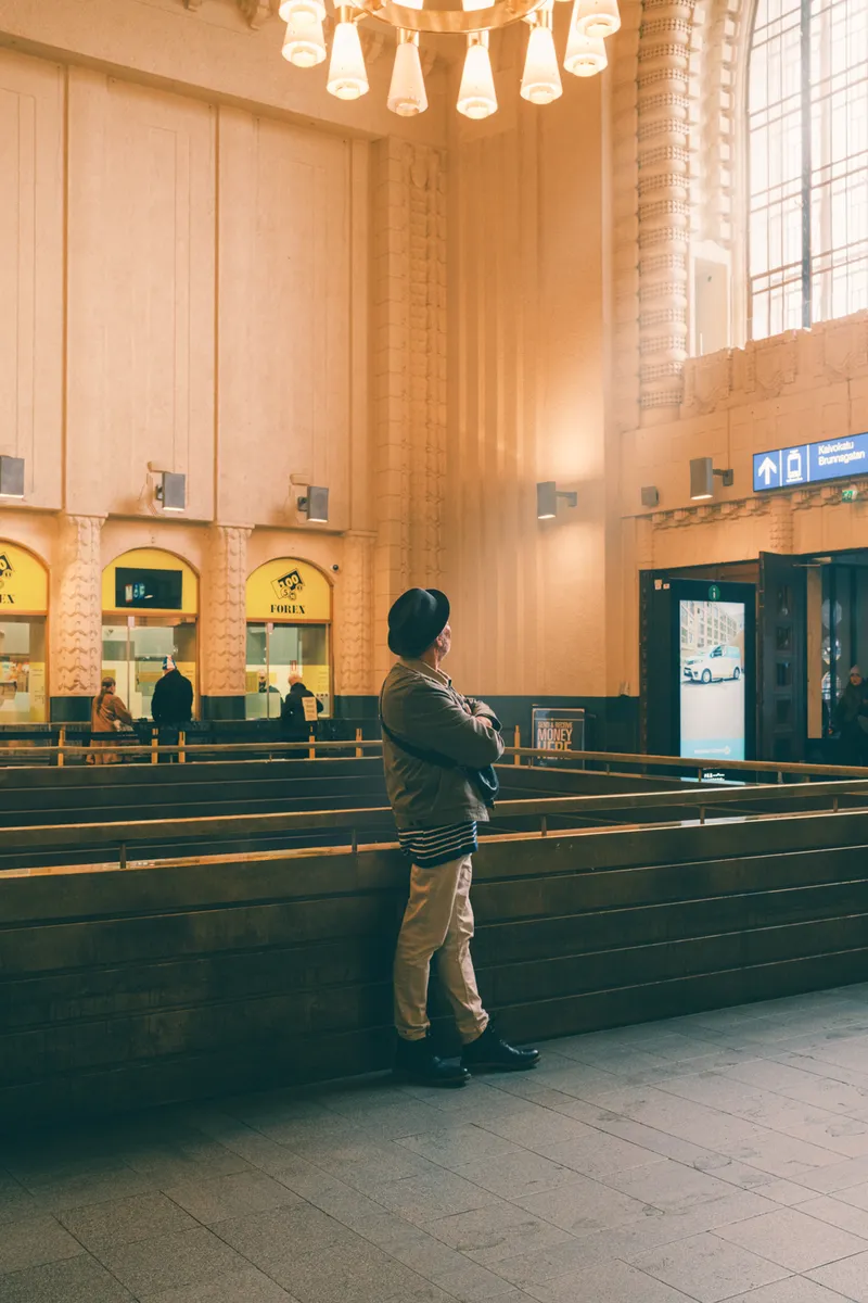 A man in a hat leans against a railing inside Helsinki Central Station, arms crossed, beneath a glowing chandelier and towering stone columns.
