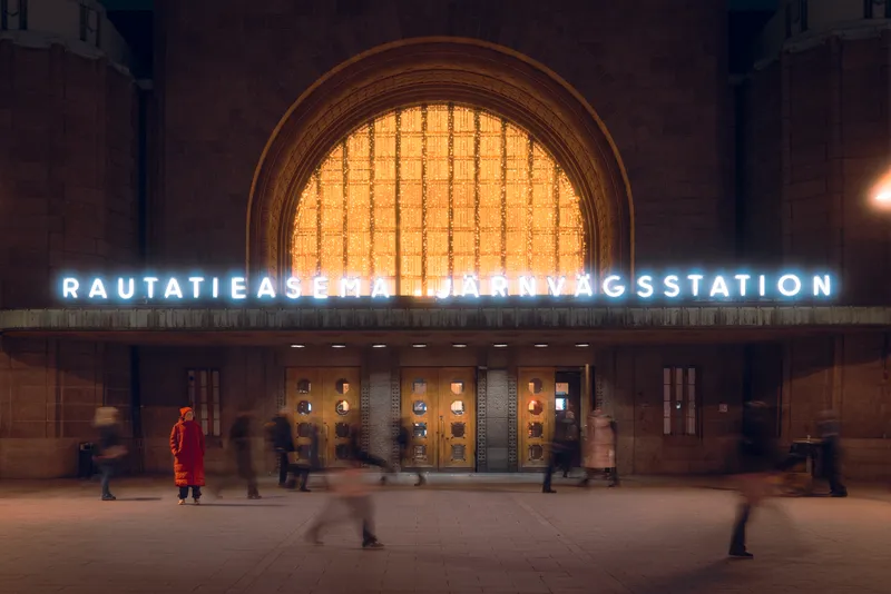 A woman in a bright orange coat stands still outside Helsinki's Rautatieasema Järnvägsstation at night, blurred figures streaming past beneath the station's illuminated sign.