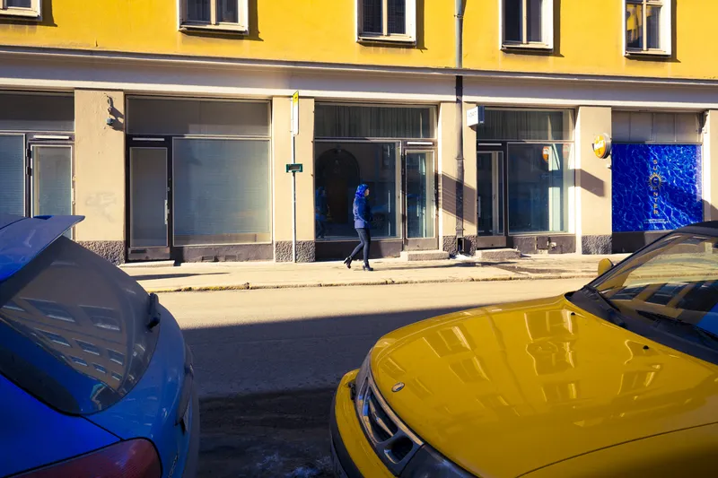 A woman in a blue coat walks past a yellow apartment building in Helsinki, a yellow car and a blue car parked at the curb below.