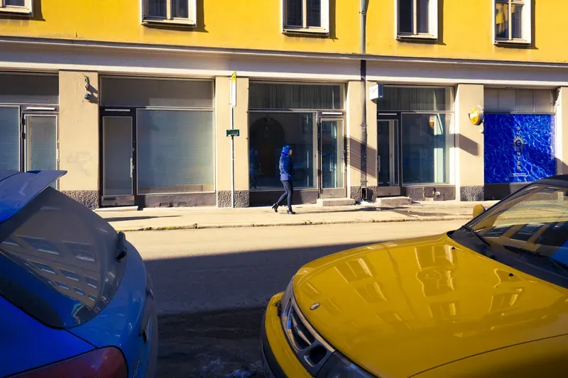A woman in a blue coat walks past a yellow apartment building in Helsinki, a yellow car and a blue car parked at the curb below.