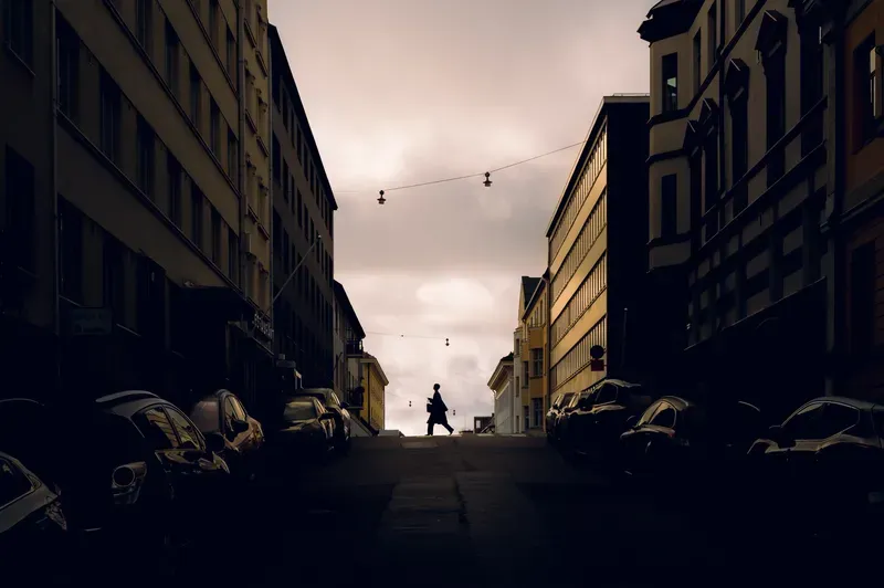 A lone figure carrying a bag crosses the crest of a street in silhouette, framed by dark buildings and parked cars under a pale pink sky strung with hanging lamps.