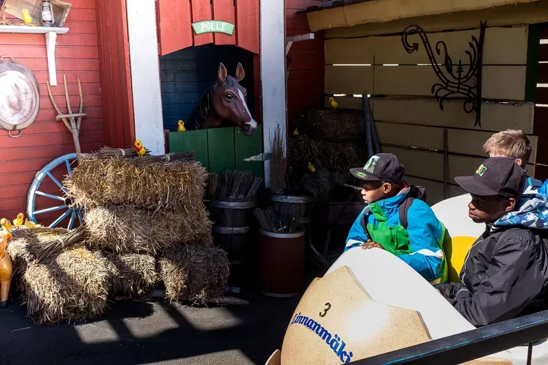 Two boys in baseball caps ride in a giant egg-shaped car in front of a painted horse stall with hay bales and toy chicks at an amusement park.