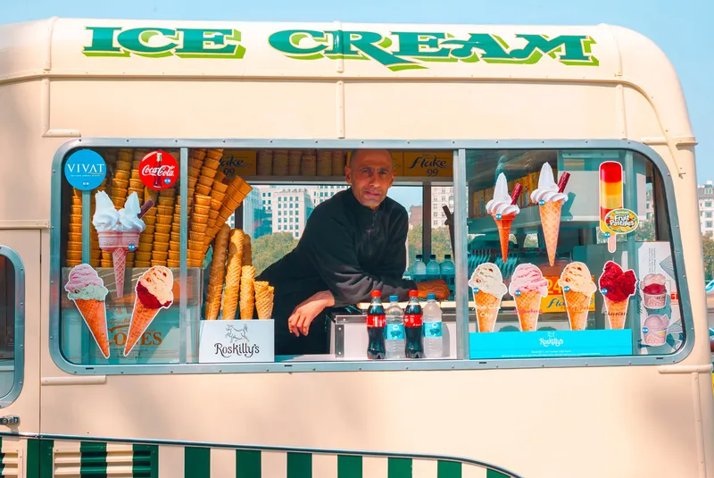 A vendor leans out of the serving window of a cream-colored Ice Cream van, cones and price cards stacked around him.