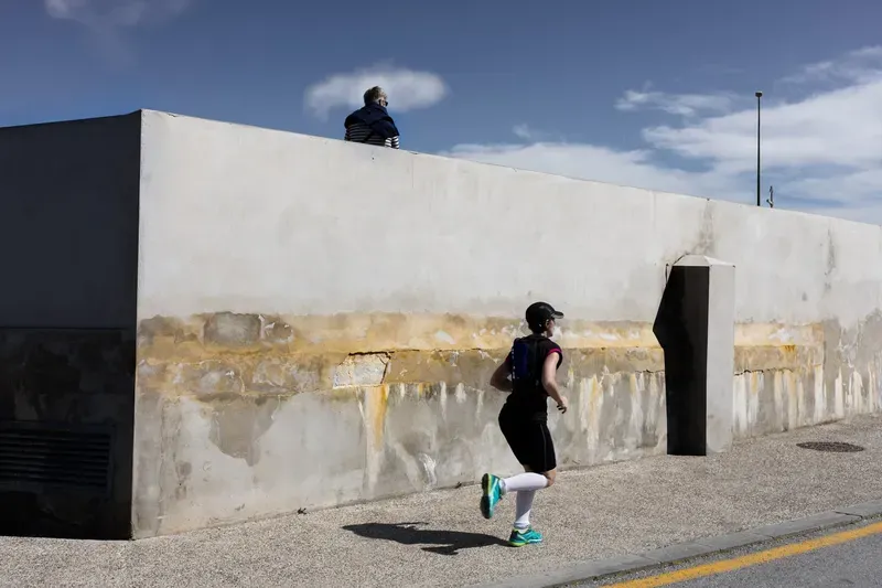 A woman jogs along a stained concrete seawall while a man sits perched on top gazing out at the sky.