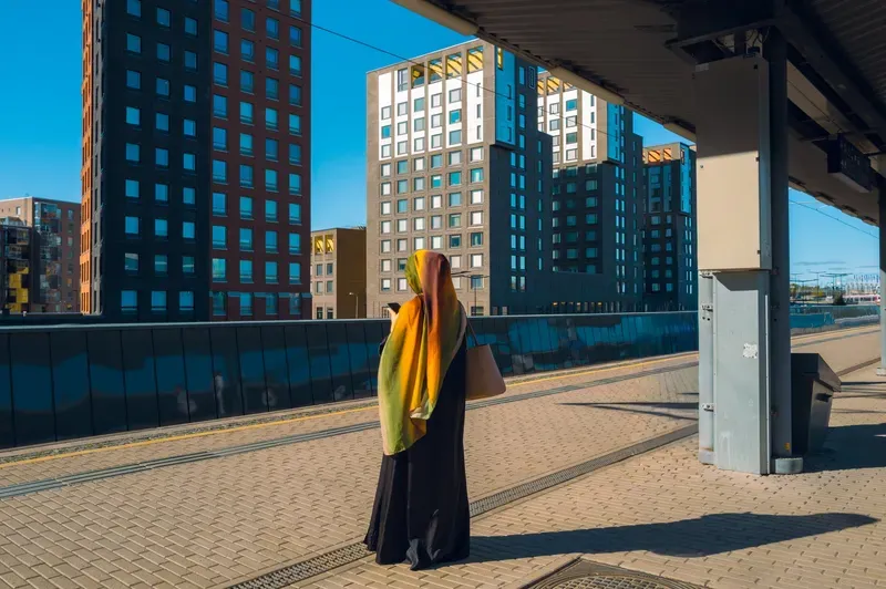 A woman in a black abaya and a green and orange headscarf reads her phone on a sunlit tram platform, new brick and glass towers rising behind her against a deep blue sky.