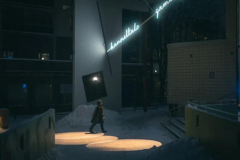 A figure walks through pools of spotlight on snow beneath a glowing cursive sign reading Kanneltalo Gamlasgården at night.