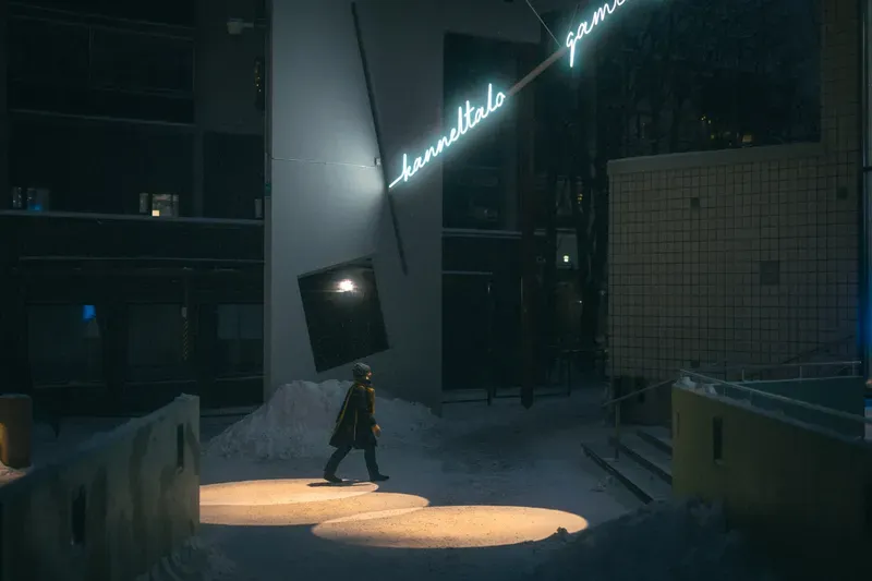 A figure walks through pools of spotlight on snow beneath a glowing cursive sign reading Kanneltalo Gamlasgården at night.