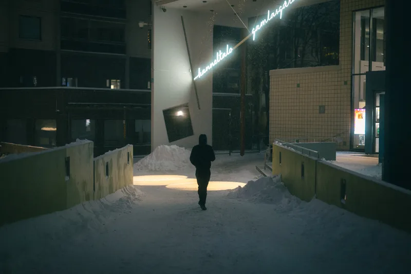 A figure in a hooded coat walks toward a yellow building past a glowing cursive neon sign on a snowy night.