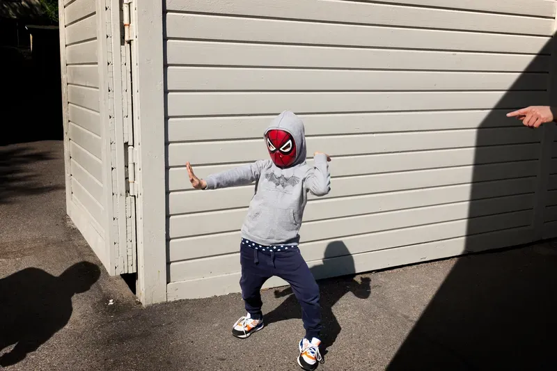 A child in a Spider-Man mask and gray hoodie strikes a fighting pose against a pale wood-paneled wall, an adult's pointing hand and both their shadows visible at the edges of the frame.