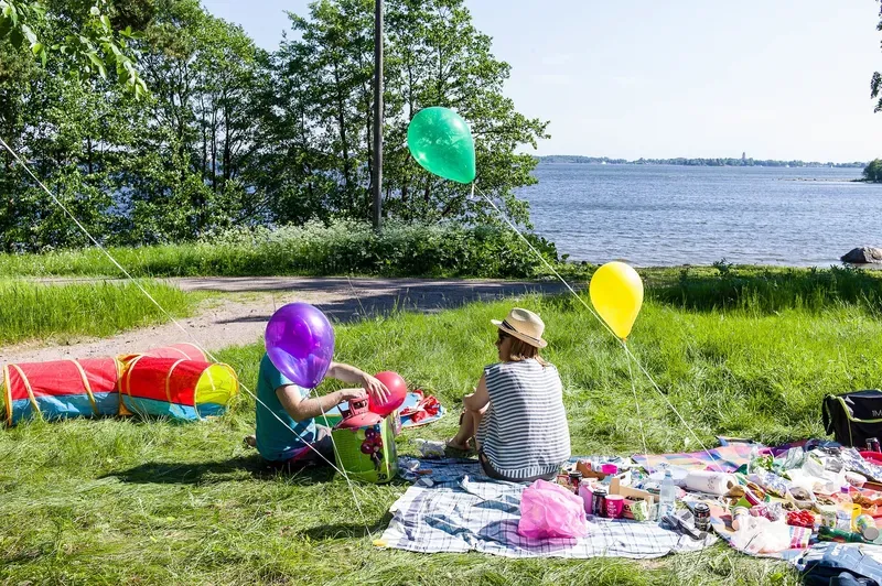 A woman in a straw hat and striped top sits on a picnic blanket by a lake beside a child filling balloons, a green and a yellow balloon floating above and a red play tunnel on the grass.