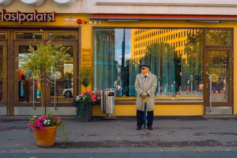 An elderly man in a pale trench coat, flat cap and sunglasses stands with a blue umbrella planted like a cane against the yellow Lasipalatsi facade, the street reflected in the window beside him.