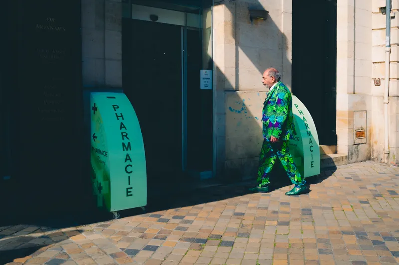 A man in a bold green tropical-print suit strolls past a curved teal pharmacy sign on a sunlit stone street.