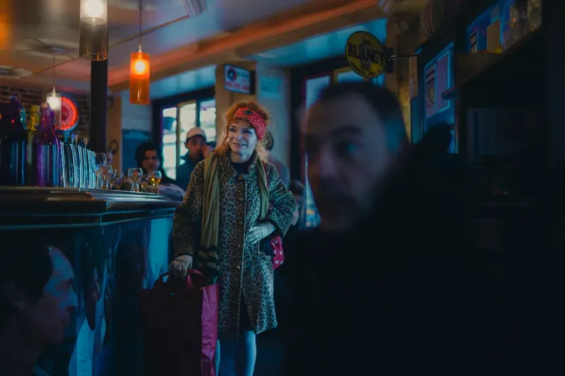 A woman in a leopard-print coat and red headscarf smiles at a candlelit bar, bags in hand, other patrons in soft focus around her.