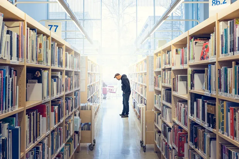 A man in a flat cap browses a library aisle, tall windows flooding the space with daylight at the far end.