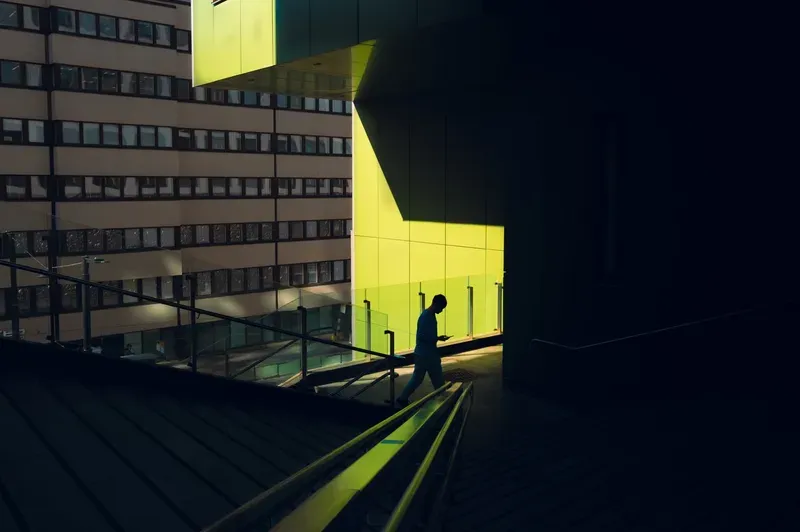 A lone figure in a striped top walks down dark stairs, head bowed over a phone, framed against a slab of sunlit chartreuse wall.