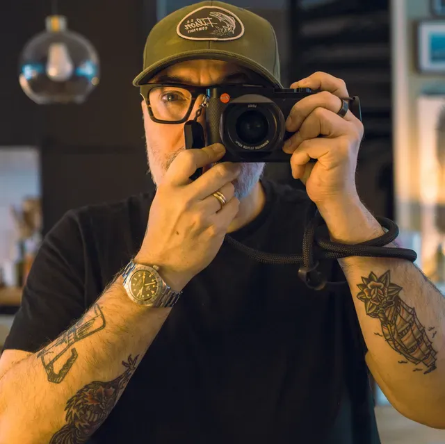 Loïc Sans holds a Leica camera up to his face, framing a self-portrait indoors.