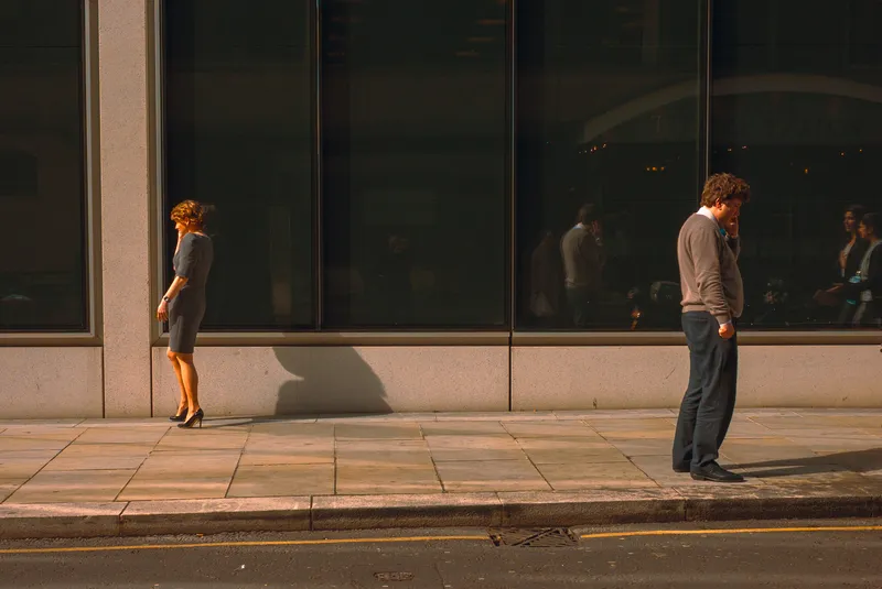 A man and woman stand apart on a sunlit sidewalk in front of dark glass office windows, both looking down at their phones.