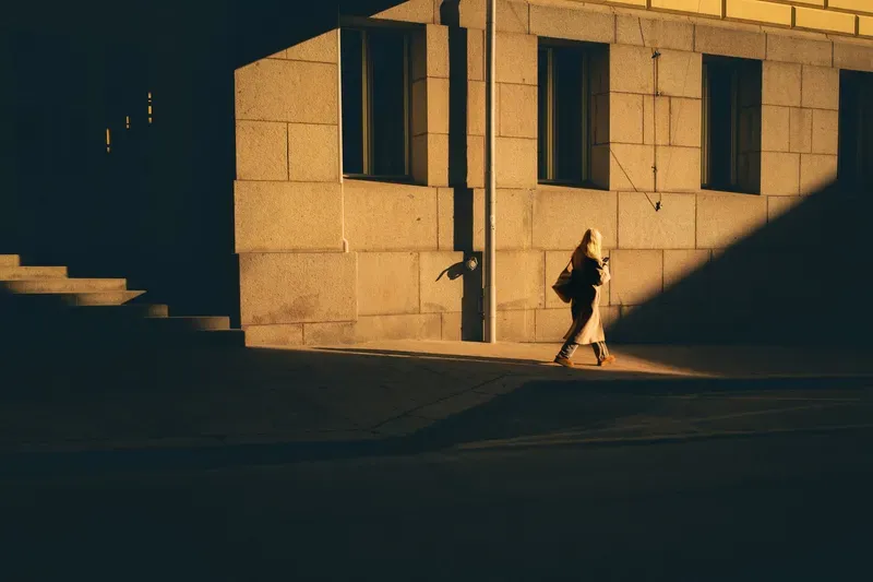 A blonde woman checks her phone while walking past a sunlit stone building corner, a sharp triangular shadow slicing across the scene.