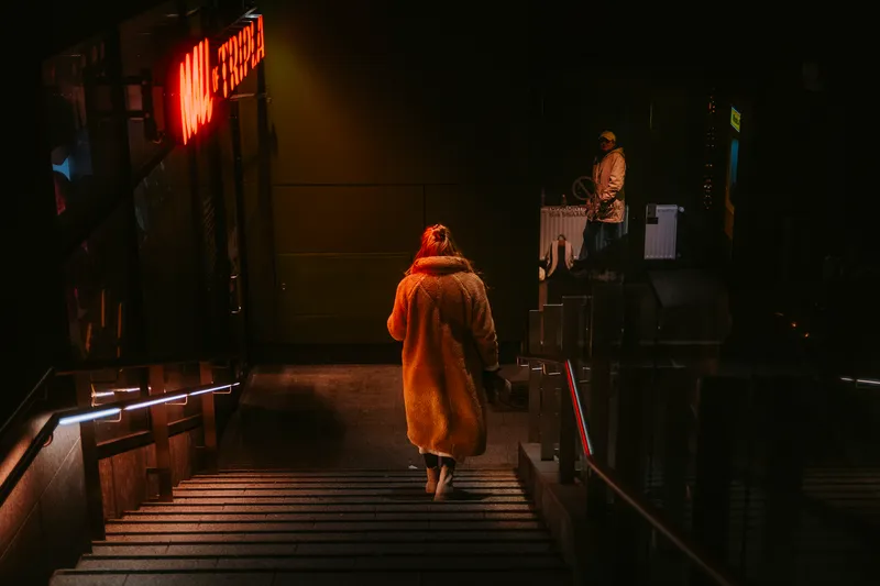 A woman in a long tan coat descends into a stairwell lit by a red Mall of Tripla neon sign, another figure waiting near the entrance behind her.
