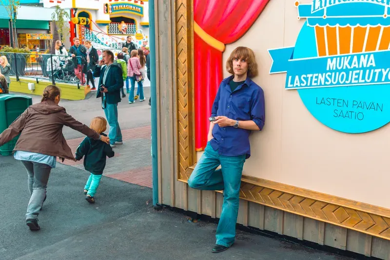 A long-haired man in a blue shirt leans against a painted wall with a red curtain and a charity sign at an amusement park, a woman hurrying a small child past behind him.
