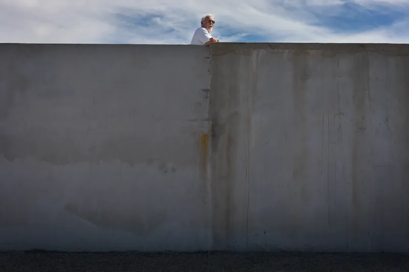An elderly man in sunglasses leans over a tall concrete wall, framed against a bright blue sky with wispy clouds.