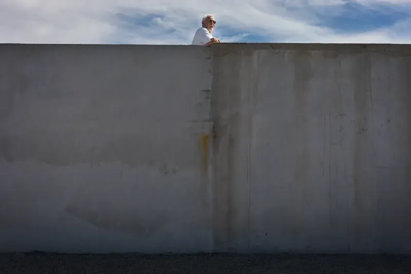 An elderly man in sunglasses leans over a tall concrete wall, framed against a bright blue sky with wispy clouds.